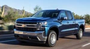 Blue 2019 Chevrolet Silverado driving on a paved road with trees, mountains, and cacti in the background