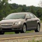 Tan 2012 Chevrolet Malibu sedan driving down a paved road past green grass and trees
