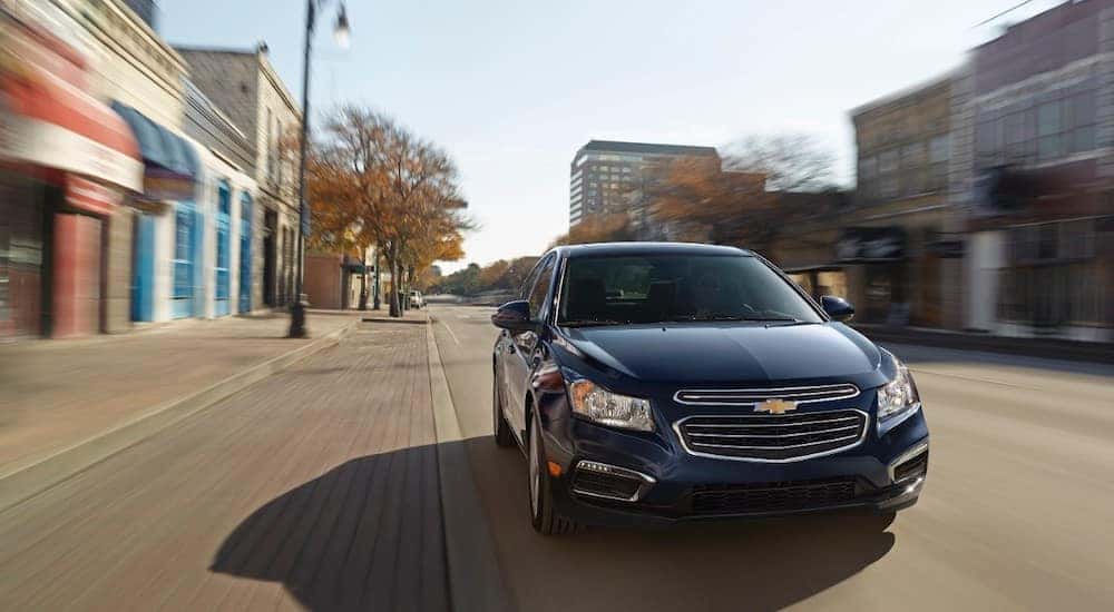 Front view of a black 2015 Chevrolet Cruze driving down the street past various storefronts