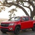 Red 2016 Chevrolet Colorado pickup truck parked next to a tall, twisty tree with fields in the background