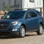 Blue 2016 Chevrolet Equinox SUV parked in a dirt lot next to a wooden fence and in front of a grey farm building
