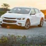 White 2016 Chevrolet Malibu parked across a paved street with mountains and sun in the background