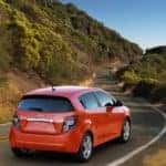Orange 2016 Chevrolet Sonic hatchback driving up a paved road by trees and tall hills