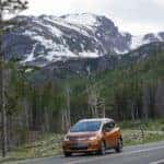 Orange 2017 Bolt EV hatchback driving down a twisty paved road in front of tall, snowy mountains
