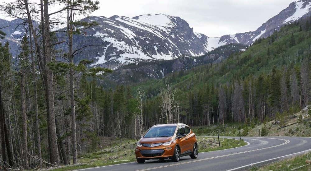 Orange 2017 Bolt EV hatchback driving down a twisty paved road in front of tall, snowy mountains