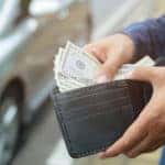 Man in a blue button-up shirt pulling cash out of a wallet next to a silver sedan