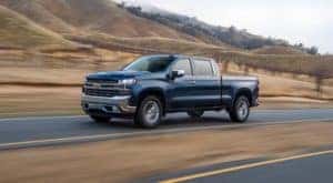 A Chevy diesel truck for sale near you, a blue 2021 Chevy Silverado 1500, is driving on a road past hills.