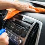 Hands holding a blue spray bottle and an orange cloth, cleaning the dashboard of a vehicle's black interior
