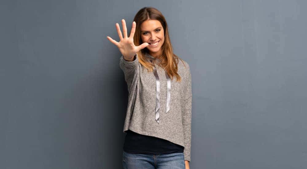 Woman in a grey sweatshirt standing against a blank wall and holding out 5 fingers