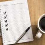 Open notebook with five checked-off boxes going down the left side, sitting next to a smartphone and a cup of coffee
