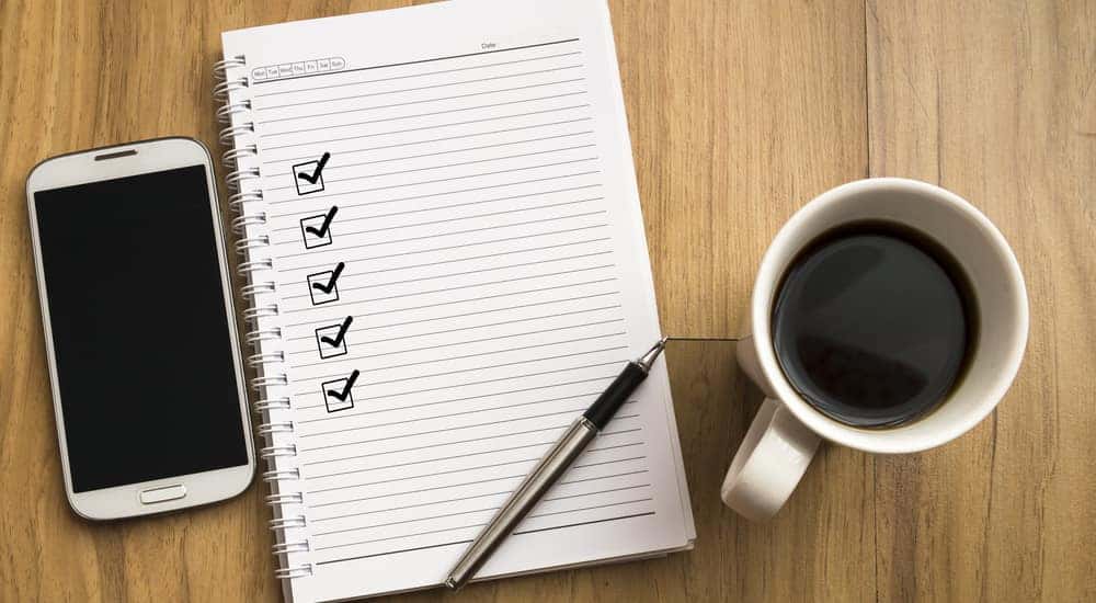 Open notebook with five checked-off boxes going down the left side, sitting next to a smartphone and a cup of coffee