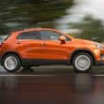 Orange 2016 Chevrolet Trax SUV driving down a paved road past green grass and trees