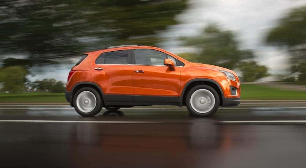 Orange 2016 Chevrolet Trax SUV driving down a paved road past green grass and trees