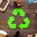 Green recycling symbol sitting on a wooden table in the middle of various paper and plastic products