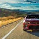 Red 2016 Chevrolet Camaro driving on a paved road past tall grass and mountains