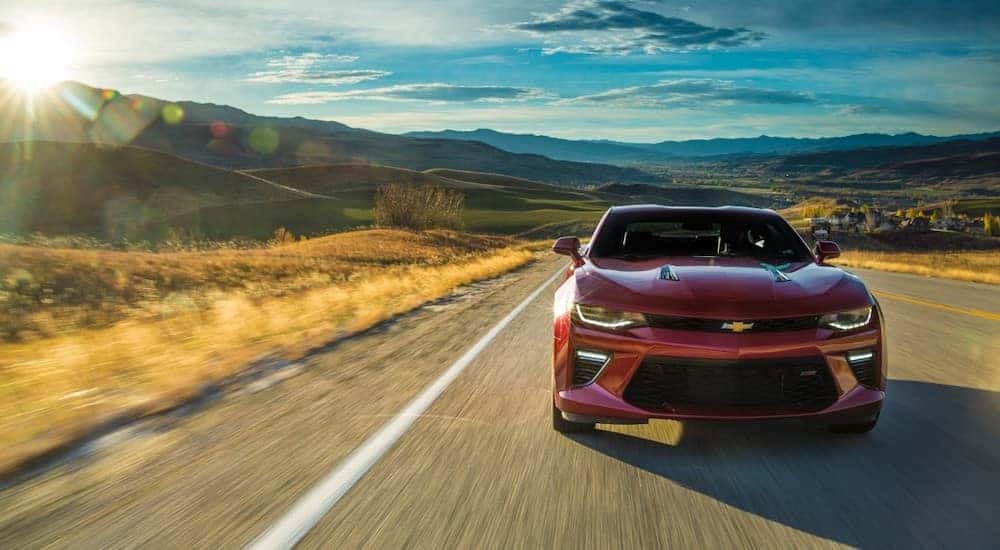 Red 2016 Chevrolet Camaro driving on a paved road past tall grass and mountains