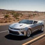 Silver 2016 Chevrolet Camaro convertible sports car driving down a paved road next to flat dirt earth