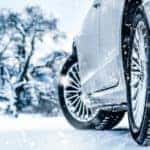 Tires on a silver vehicle driving down a snowy road past snow-covered trees
