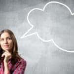 Woman in a red shirt with her hand on her chin standing in front of a grey wall with a white "thinking" bubble drawn on it