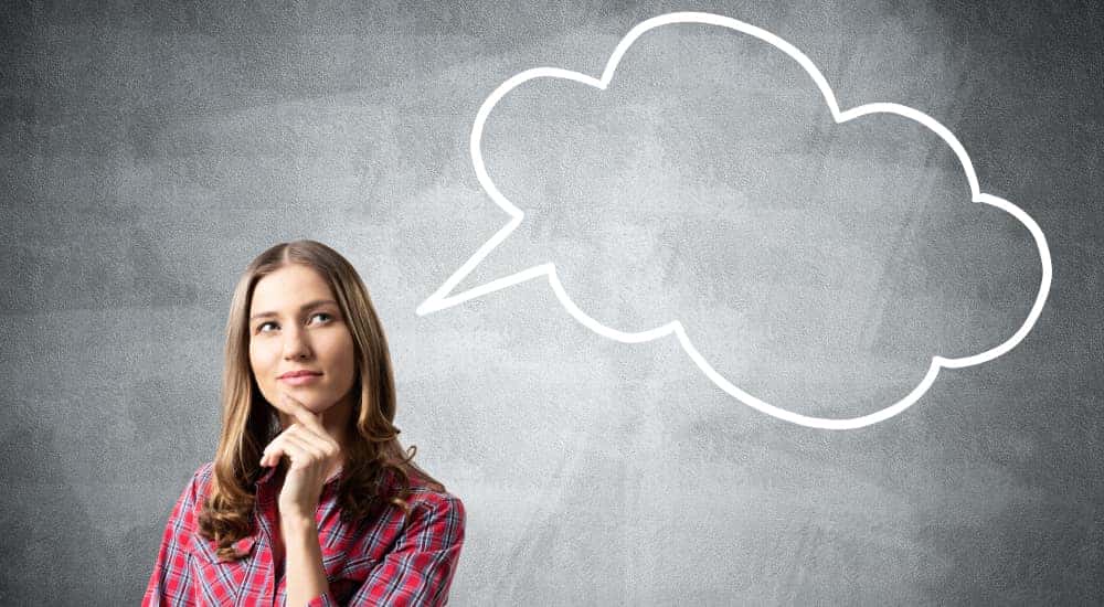 Woman in a red shirt with her hand on her chin standing in front of a grey wall with a white "thinking" bubble drawn on it