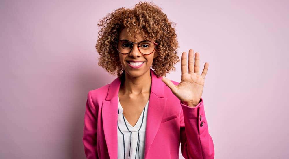 Woman in a pink blazer standing with five fingers up in front of a pink wall