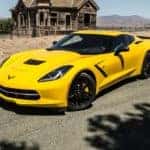 Yellow and black 2016 Chevrolet Corvette sports car parked on a paved road in front of sand and a small brown building