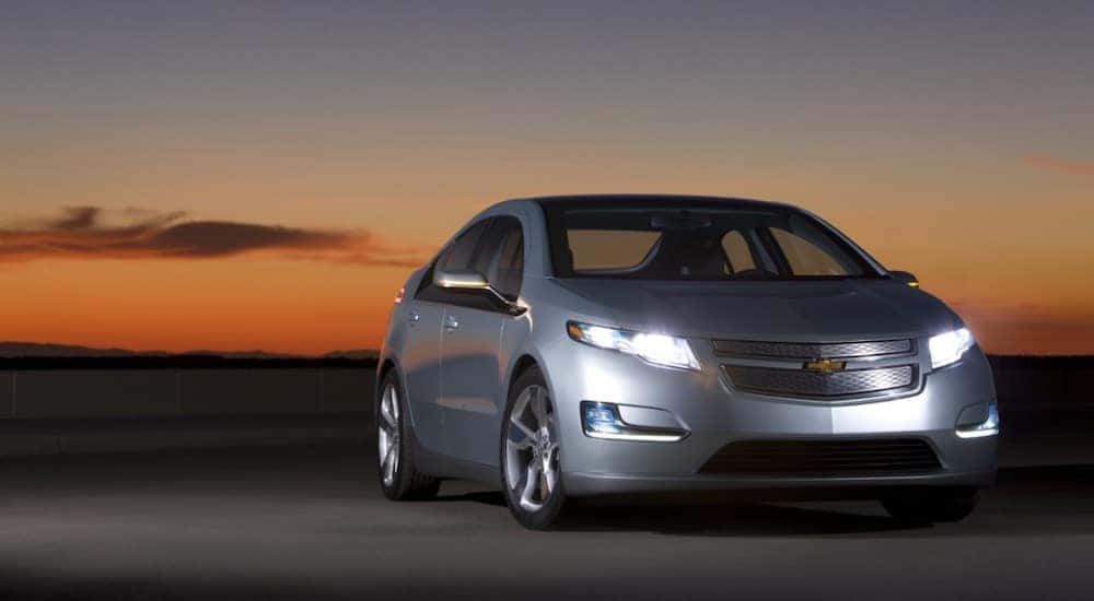 Grey 2011 Chevrolet Volt with its lights on parked in front of a dark sunset
