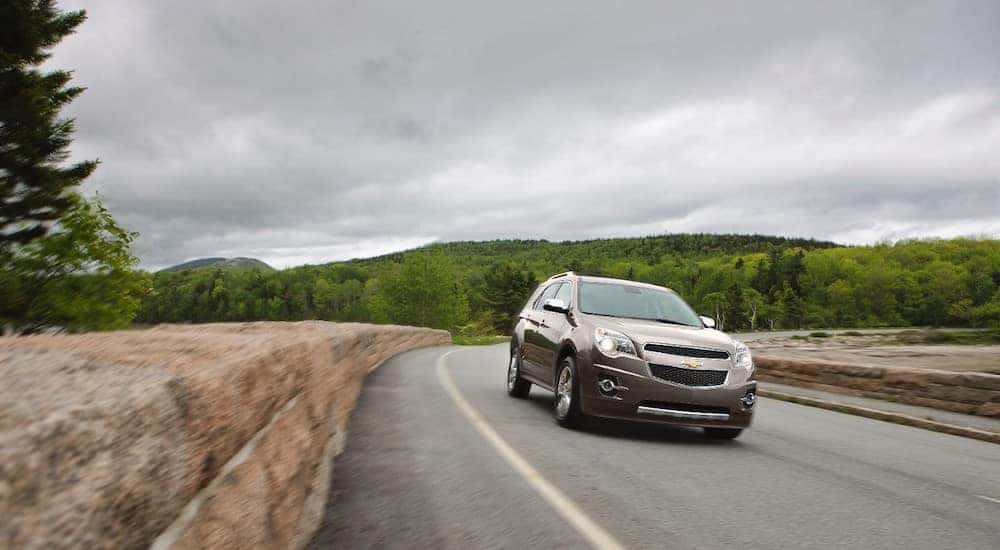 Tan 2015 Chevrolet Equinox SUV driving down a paved road past a stone wall