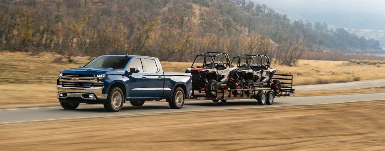 A blue 2021 Chevy 1500 diesel is towing side-by-sides past yellow grass and a hill.