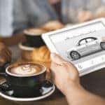 Hands holding a tablet with a car displayed on the screen above a table with coffees on it