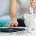 Hands holding cash and using a calculator on a white table next to a white piggybank