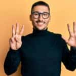Man in a black turtle neck and glasses holding up eight fingers against an orange wall