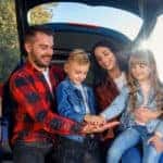 Mom and dad in plaid shirts holding a son and daughter in jean jackets while they sit in the open trunk of a vehicle