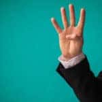Hand of a man in a suit holding up four fingers in front of a blue-green wall