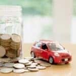Pile of coins next to a jar of coins and a red toy car on a wooden surface