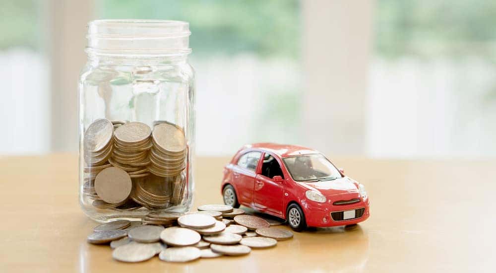 Pile of coins next to a jar of coins and a red toy car on a wooden surface