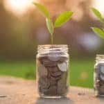 Two jars of coins with plants sticking out of the top sitting on a table outside in front of grass