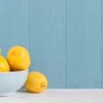 Yellow lemons in a white bowl sitting on a table in front of a blue wall