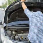 Man in a plaid shirt looking under the open hood of his car on the side of the road
