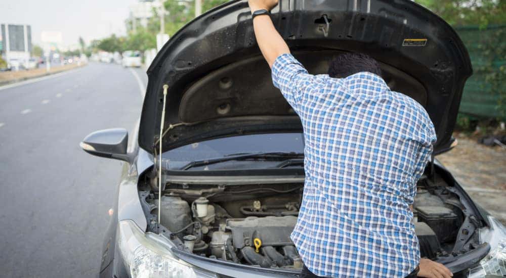 Man in a plaid shirt looking under the open hood of his car on the side of the road