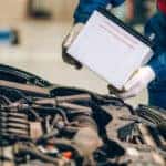 Male mechanic in a blue and red jumpsuit placing a white car battery under the open hood of a vehicle