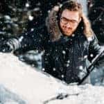 Man in a black winter jacket clearing snow off of the windshield of his vehicle