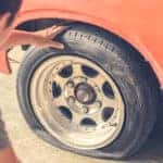 Man in a t-shirt crouching down to inspect the flat tire on an orange car