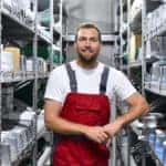 Man in a white shirt and red overalls standing between tall shelves of auto parts