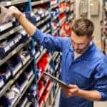 Mechanic in a blue jumpsuit grabbing car parts off of a shelf while looking at a tablet