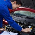Mechanic in a blue jumpsuit looking under the hood of a red vehicle with a clipboard