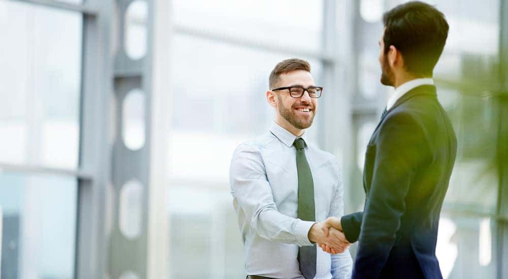 Man in a suit shaking hands with a man in a button-up and tie in front of a glass building