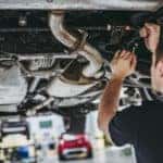Two men, wearing all black, working on a car that is lifted in a car shop