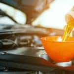 Hands pouring oil from a grey bottle into the oil tank of a vehicle through an orange funnel