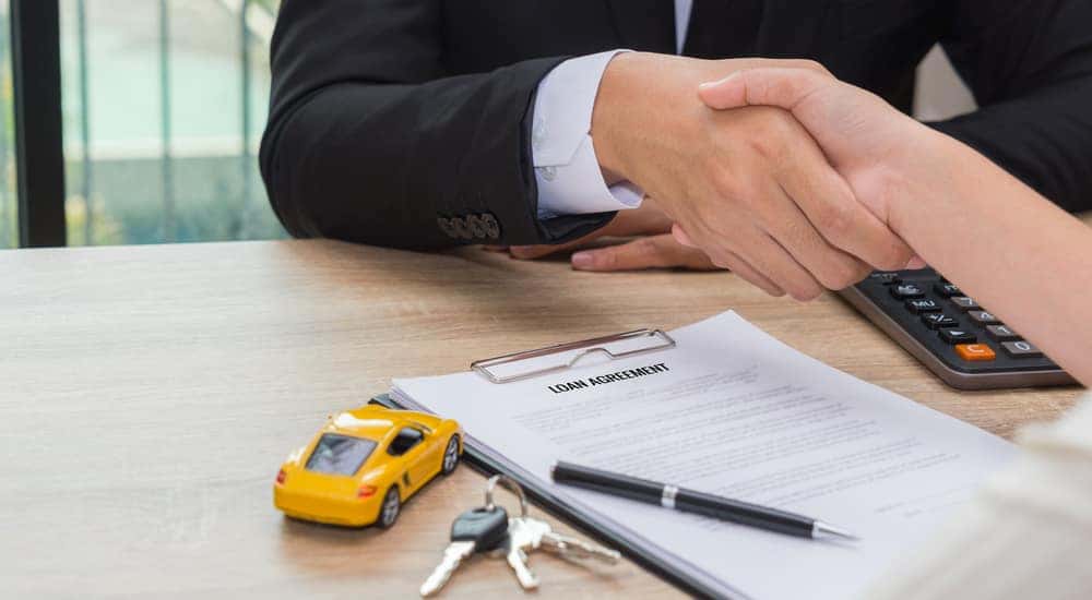 Two people shaking hands over a brown desk with a loan, pen, calculator, toy car, and car keys on it